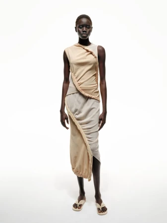 Full-length studio shot of an adult female fashion model with dark skin, posing in a draped beige dress and matching flip-flops.