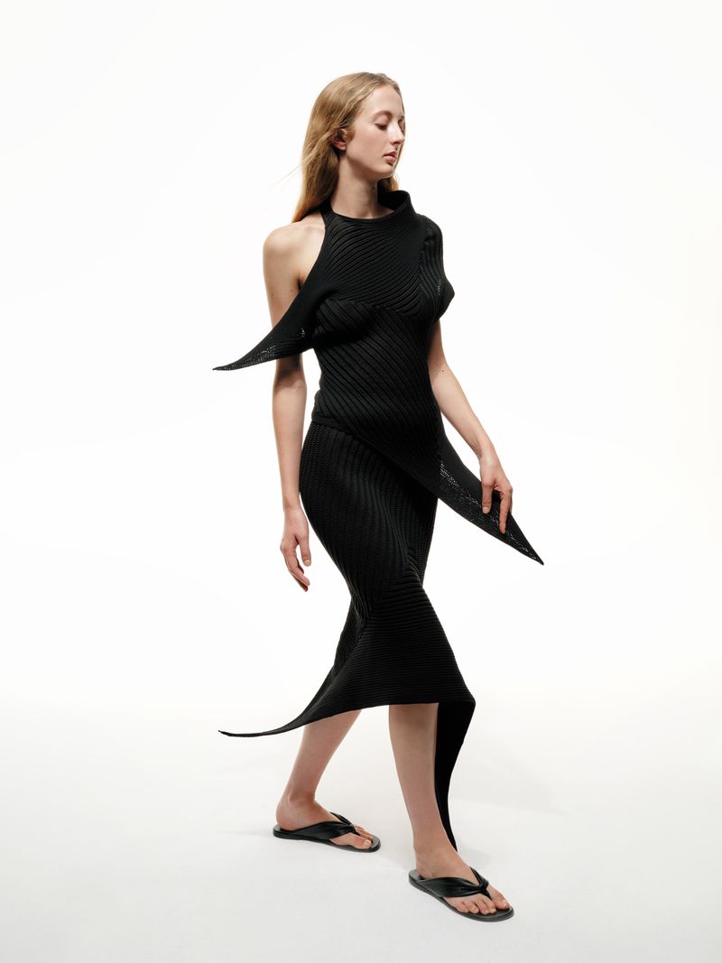 A slender woman with fair hair walks in a black ribbed halter dress and black thong sandals against a bright white background.