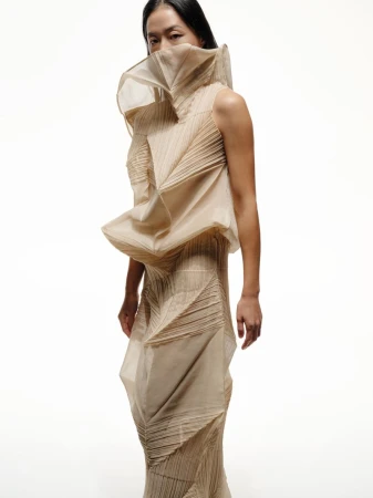 Woman poses in a studio wearing a sculptural beige sleeveless top and long skirt, with a unique folded and textured design.