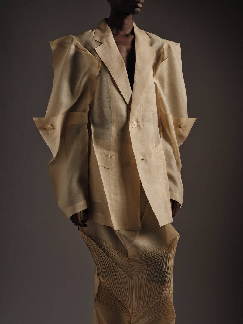 Fashion shot of a Black model in beige linen suit with exaggerated shoulders, posing against a neutral gray backdrop in studio lighting.