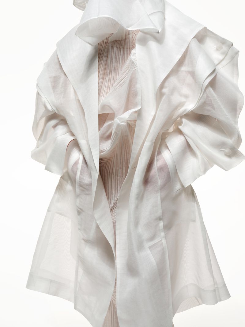 Figure wearing a sculptural white gown and coat with draping and structured forms against a bright white seamless backdrop in a studio.