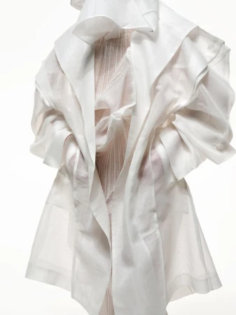 Figure wearing a sculptural white gown and coat with draping and structured forms against a bright white seamless backdrop in a studio.