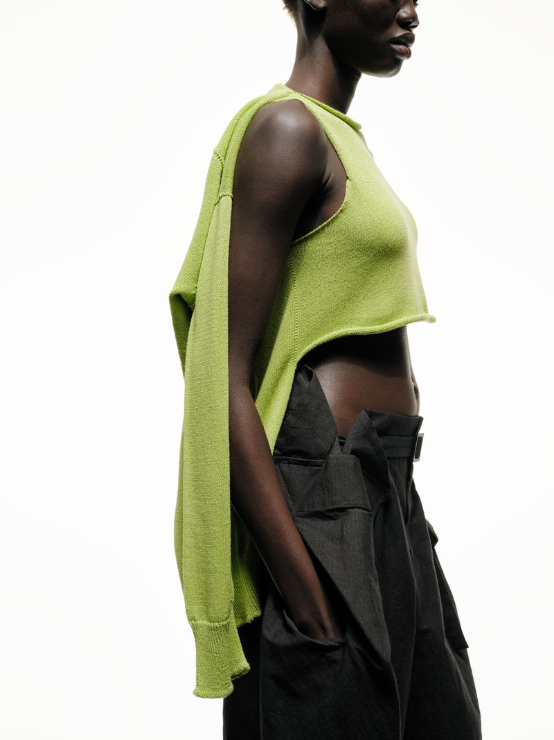 Black woman in a studio shot wearing a contemporary asymmetrical sleeveless green top and wide-leg, belted black pants against a white backdrop.