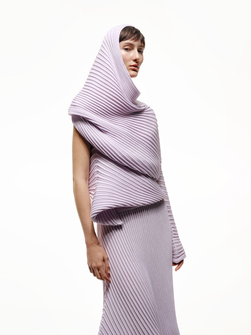 A woman in a light purple pleated garment with a hood stands against a white backdrop, showcasing an avant-garde fashion statement.
