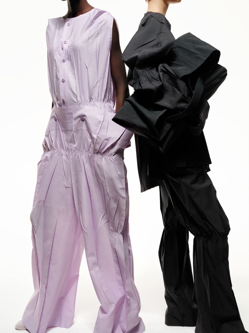 Two fashion models showcase designer jumpsuits in a studio setting, one in light purple and the other in architectural black.