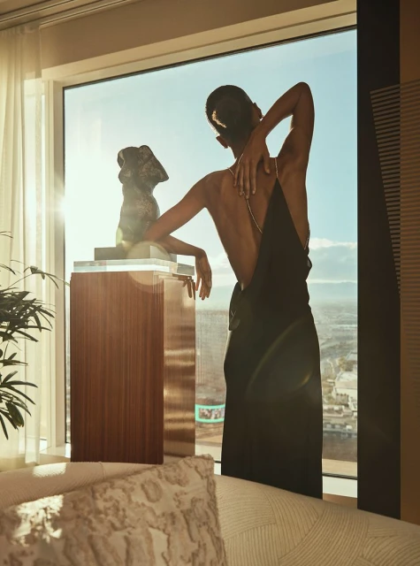 Elegant woman in black dress poses near a sculpture indoors by a window overlooking an urban landscape with bright, natural light.