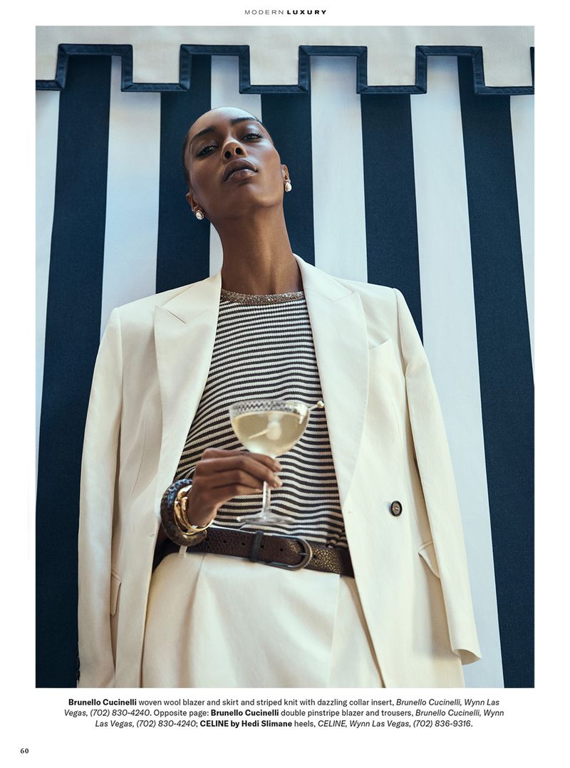Elegant woman poses in cream blazer and striped top holding drink, against a striped wall. Sophisticated fashion photography.