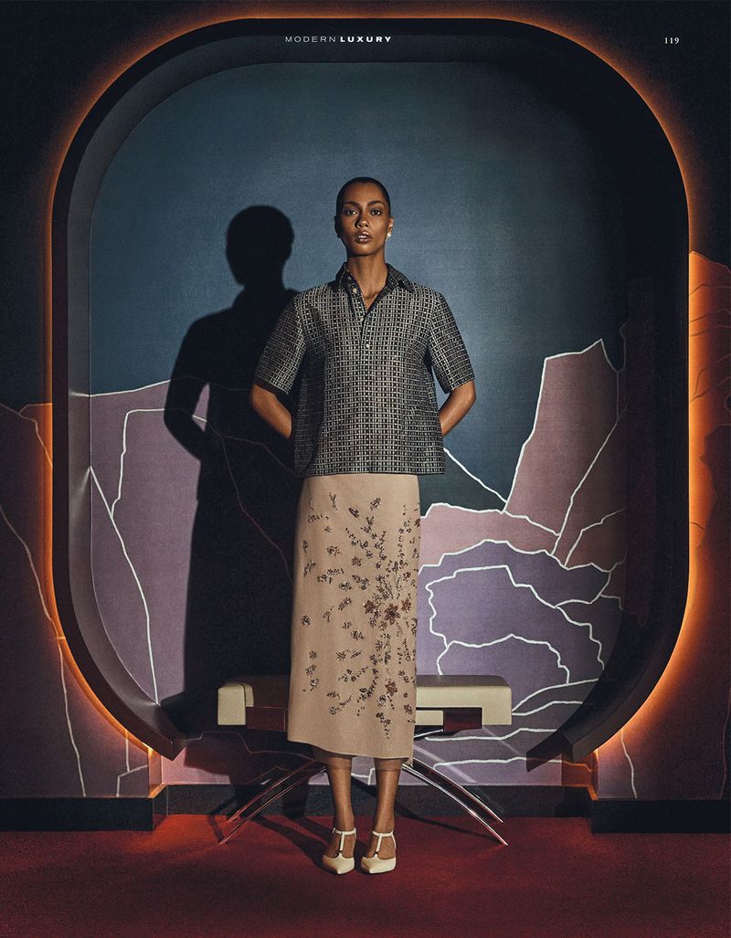 A full-length portrait of a woman in a blouse and skirt, standing in front of an archway with colorful architectural details and lighting.