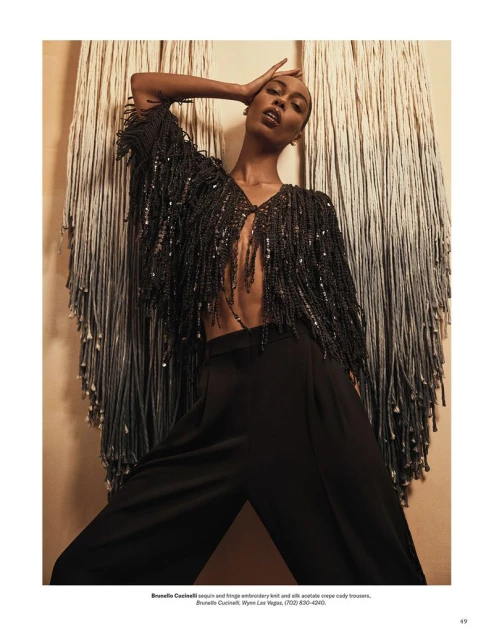 Fashion shot: A Black female model poses in a beaded fringe top and black pants, against a fringe wall hanging in warm lighting.