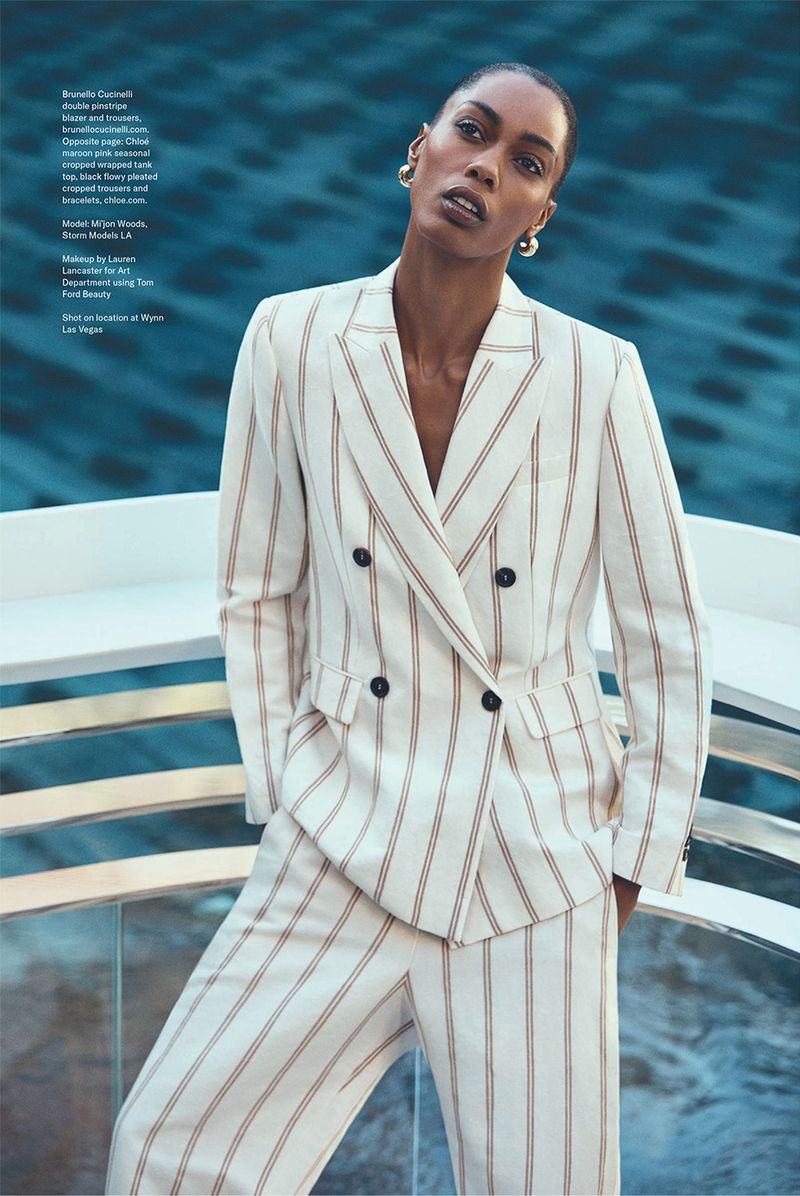 An adult female model poses in a white striped suit outdoors near an aqua background, showcasing fashion and beauty with modern elegance.