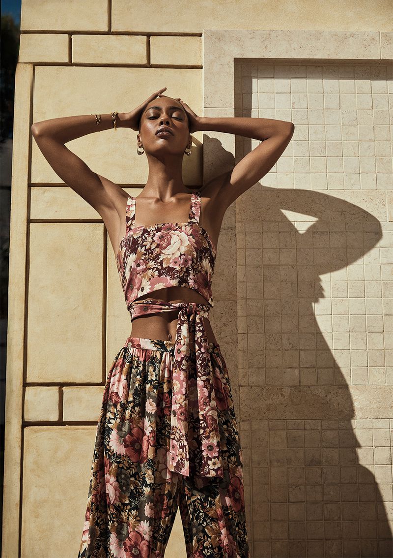 An adult female model poses in a floral-print two-piece set against a textured wall, hands on her head, jewelry, natural light.