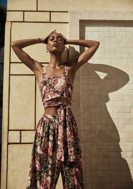 An adult female model poses in a floral-print two-piece set against a textured wall, hands on her head, jewelry, natural light.