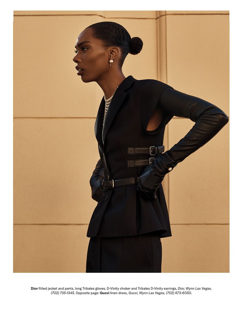 Elegant Black female model in a dark tailored jacket, pearl jewelry, and gloves against a neutral wall. Fashion editorial shot.