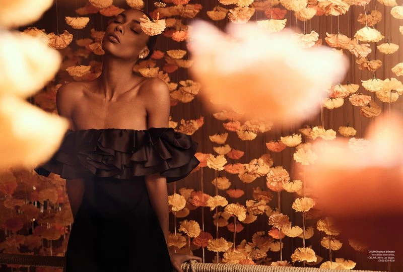 Elegant woman in a black off-the-shoulder dress poses against a backdrop of suspended yellow flowers in a fashion editorial shot.