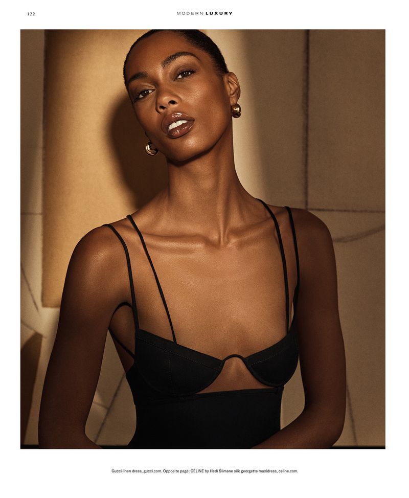 Glamorous Black woman in a black dress with delicate straps, adorned with gold earrings, poses in a warm-toned studio with soft lighting.