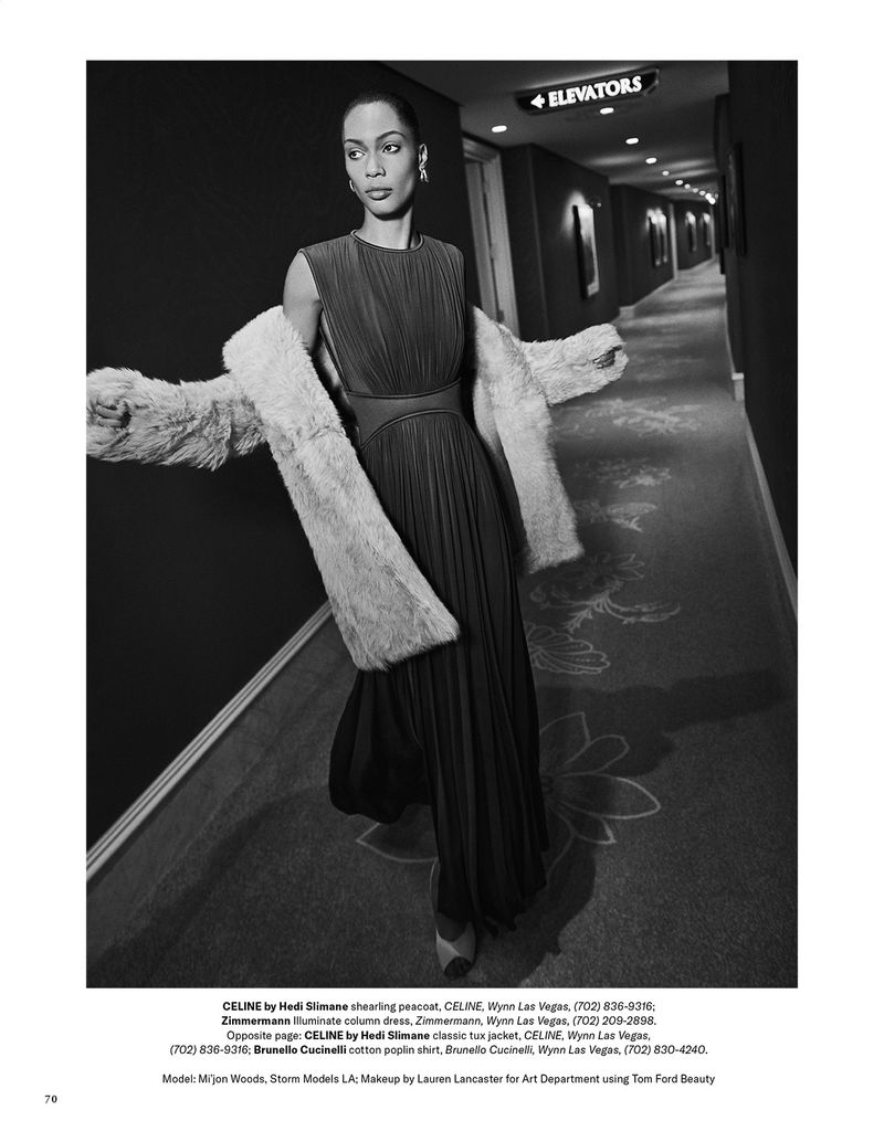 Monochrome editorial fashion shot of a woman in a pleated dress and fur coat, walking in a hotel hallway with an elevator sign.