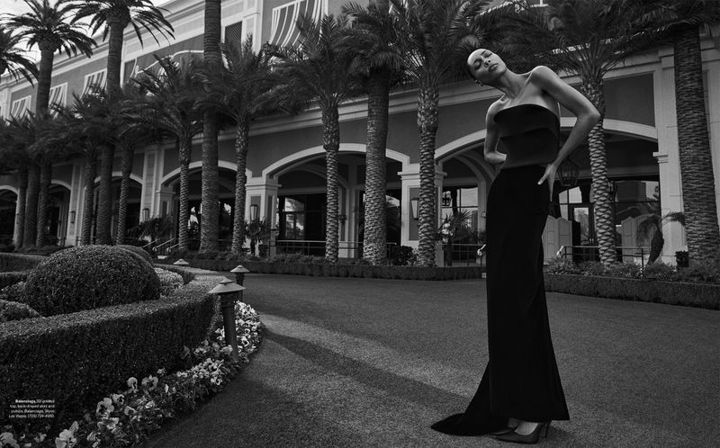 Elegant adult woman poses in strapless black dress, walking in front of resort with palm trees. Fashion editorial photography.