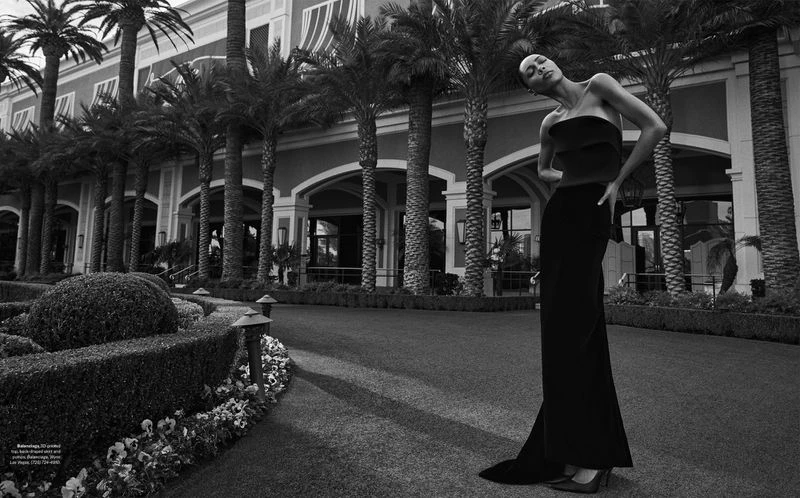 Elegant adult woman poses in strapless black dress, walking in front of resort with palm trees. Fashion editorial photography.