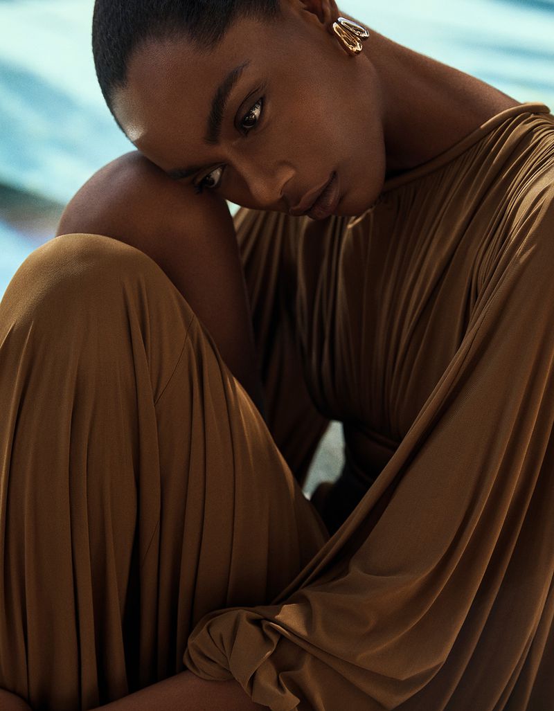 Portrait of a pensive adult woman with dark skin and gold earrings, draped in a flowing brown dress, set against a blurred backdrop.