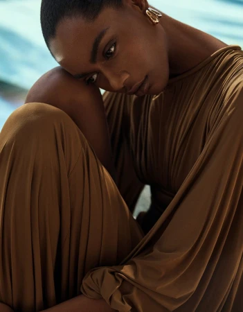 Portrait of a pensive adult woman with dark skin and gold earrings, draped in a flowing brown dress, set against a blurred backdrop.