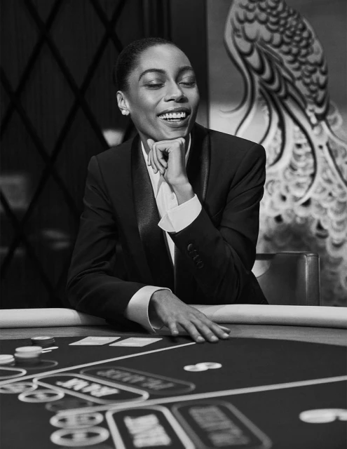 Elegant woman in a tuxedo jacket smiles at a poker table with chips and cards, indoor casino setting, black and white.