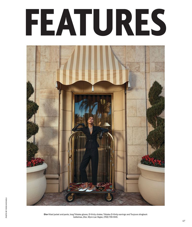 Fashion editorial featuring a woman in a black suit leaning against a luggage trolley outside a building with a striped awning.