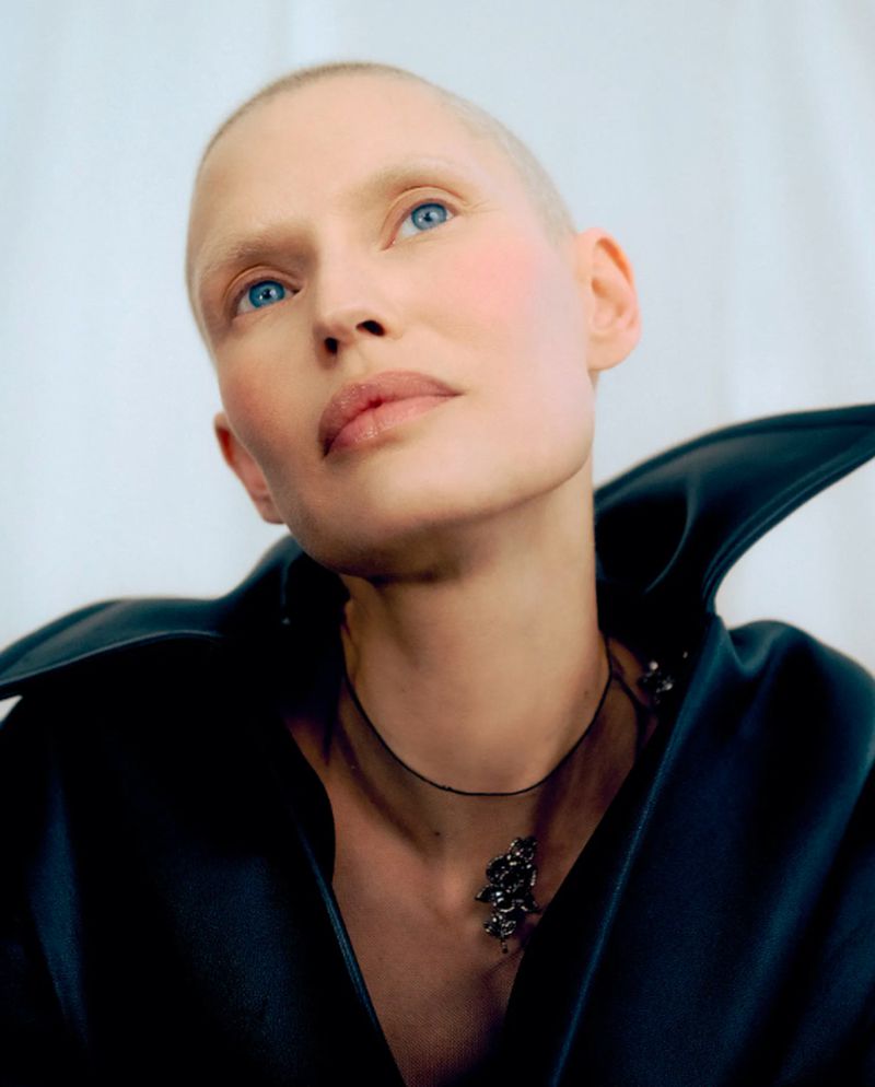 A close-up studio portrait of a fair-skinned, bald adult woman with blue eyes, wearing a black collar and necklace against a white backdrop.