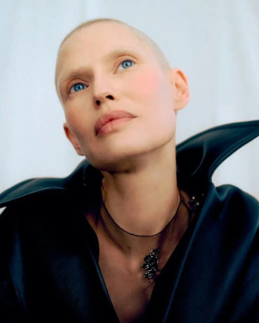 Close-up portrait of model Bianca Balti with a shaved head wearing a dark structured collar against a white background