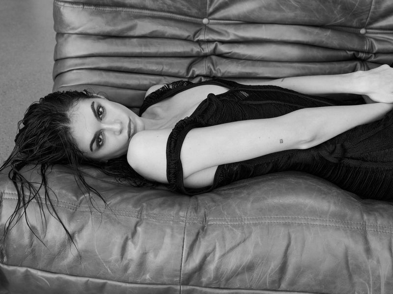 Monochrome portrait of a woman with wet hair and a dark dress reclining on a leather couch, looking directly at the camera.