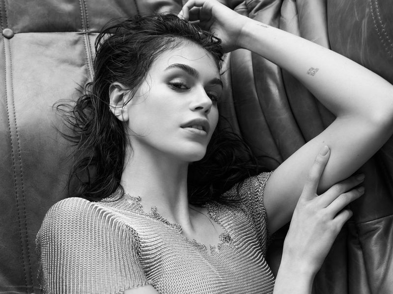 Black and white portrait of a woman lying on a leather sofa, wearing a metal mesh top and looking directly at the camera in soft light.