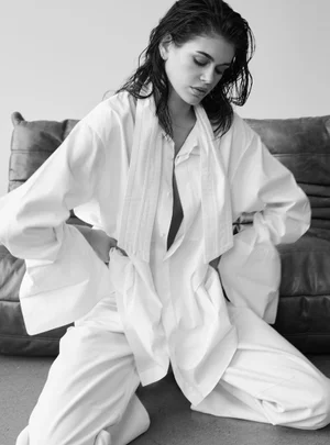 Model with wet hair wearing an oversized white shirt and trousers kneeling on a floor in front of a dark leather sofa.