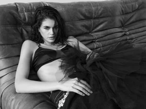 Model Kaia Gerber with wet hair in a black bralette and tulle skirt lounging on a dark leather couch.