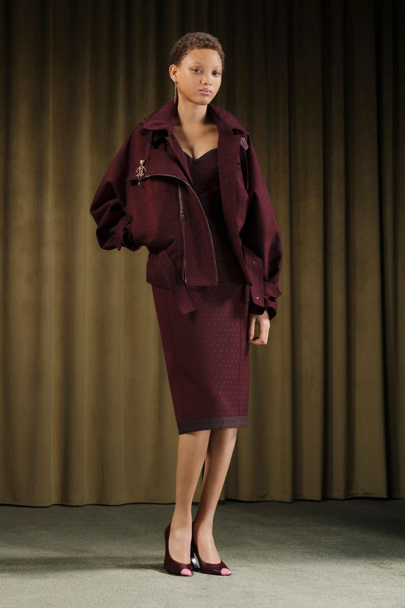 A female model in a burgundy-colored skirt suit with a motorcycle jacket poses indoors in a studio with a neutral curtain backdrop.