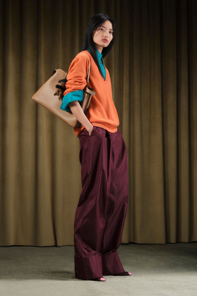 Full-length studio shot of an Asian woman wearing orange sweater, teal undershirt, burgundy wide-leg trousers, and carrying a tan handbag.