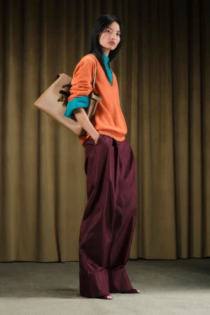 Full-length studio shot of an Asian woman wearing orange sweater, teal undershirt, burgundy wide-leg trousers, and carrying a tan handbag.