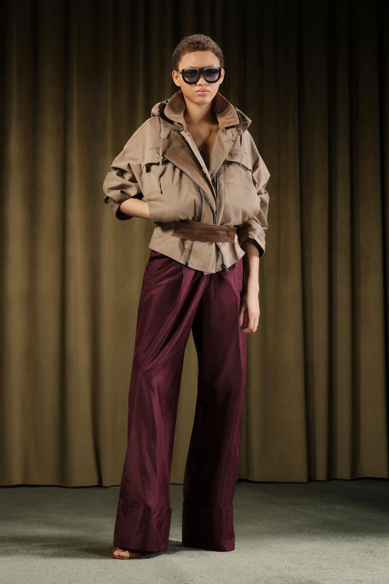 Full shot of a young adult fashion model in a tan jacket and maroon satin pants standing against a tan curtain backdrop.