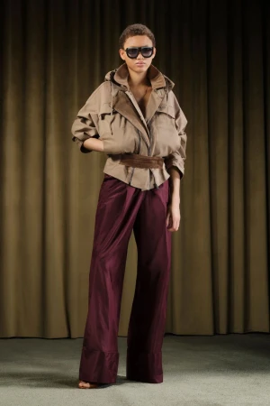 Full shot of a young adult fashion model in a tan jacket and maroon satin pants standing against a tan curtain backdrop.