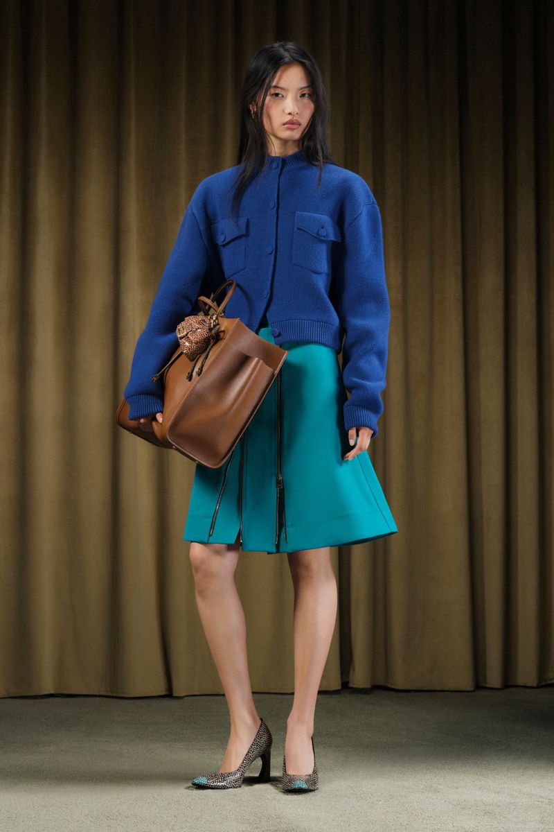 Full-length fashion shot of a woman wearing a blue jacket, green skirt, and patterned pumps while holding a brown leather tote bag.