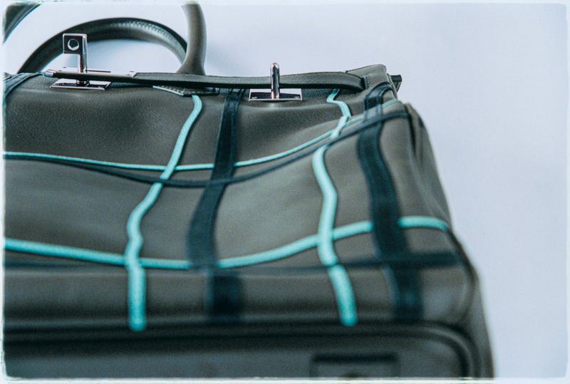 Fashionable olive-green leather handbag with blue accents, silver hardware, and a top handle on a clean white backdrop.