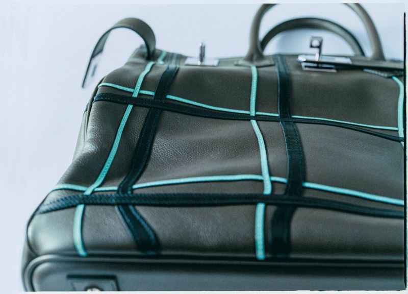 Close-up of a structured grey leather handbag with turquoise piping, with handles and silver hardware, against a white backdrop.