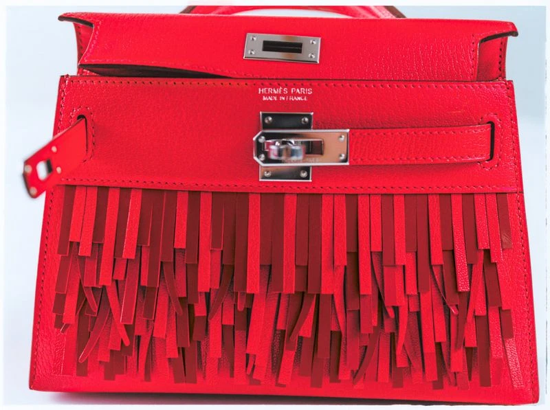 Close-up of a vibrant red leather handbag with fringe detail and silver hardware, set against a stark white backdrop in a studio setting.