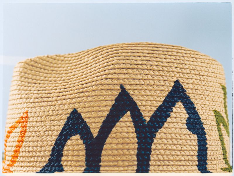 Close-up of a beige pouf with dark blue, green and orange geometric shapes knitted into the exterior material, against a light blue background.