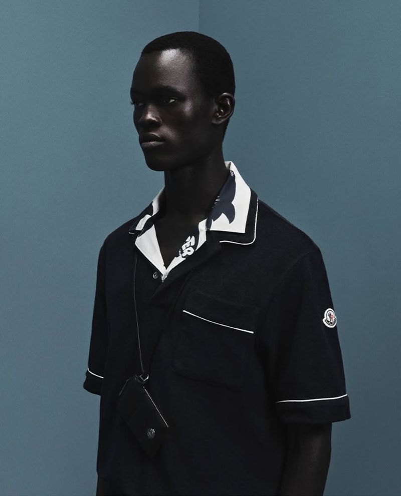 Dark-skinned male model in a black pajama-style shirt and small black cross-body bag poses against a light blue studio backdrop.