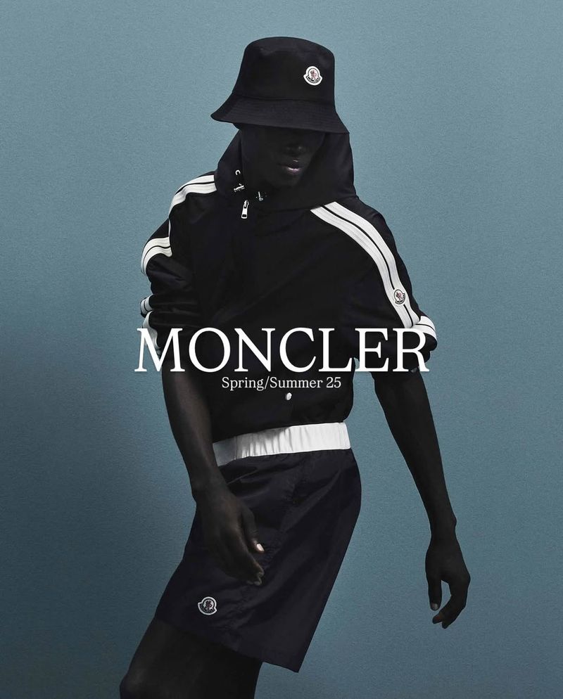 Moncler Spring/Summer 25 fashion visual features an adult male wearing a black bucket hat, jacket with white stripes, and shorts against a blue background.