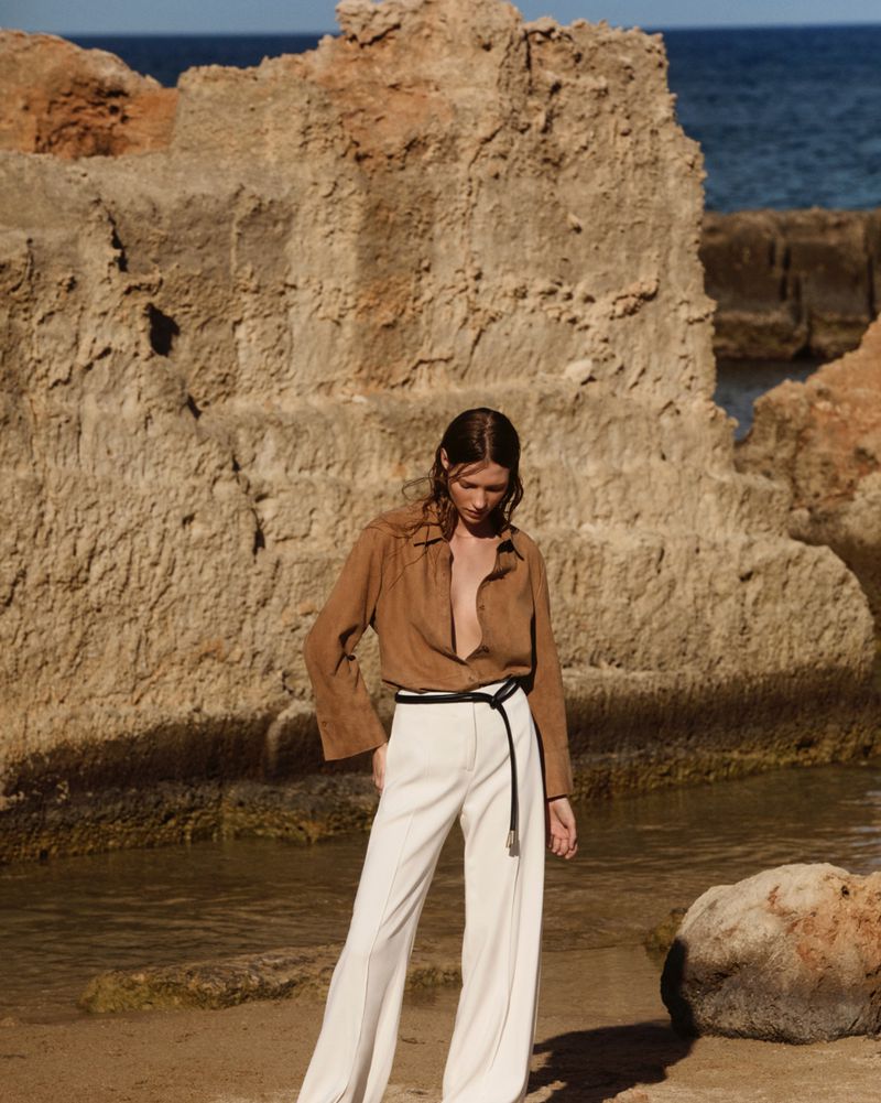 A stylish adult female poses outdoors on a sandy beach near rocky formations, wearing a brown button-up shirt and white slacks.
