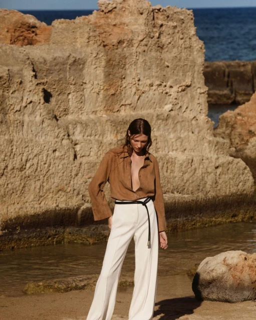 A stylish adult female poses outdoors on a sandy beach near rocky formations, wearing a brown button-up shirt and white slacks.