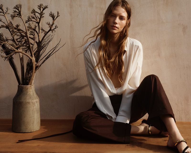 A fair-skinned woman with long wavy hair in a relaxed pose, wearing a white blouse and brown pants, sits indoors near a vase of branches.