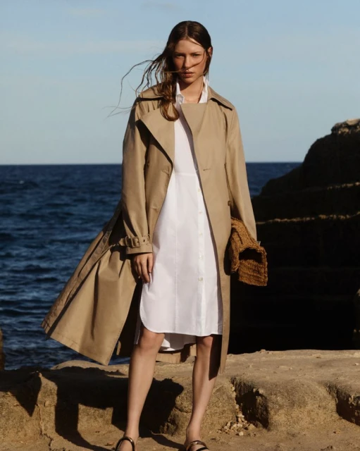 A poised woman in a tan trench coat and white dress stands on rocks by the sea, her gaze directed confidently forward in a fashion editorial.