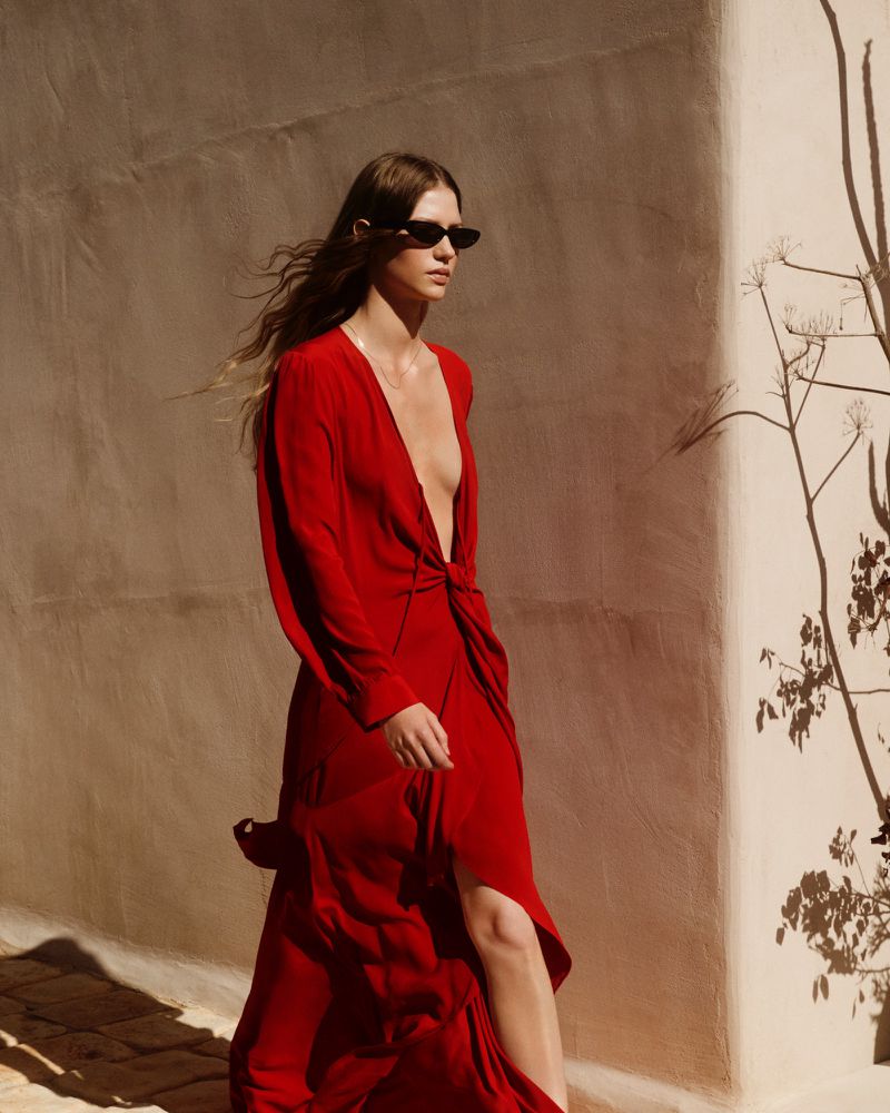 Woman in a red dress walks along a textured wall in bright sunlight, wearing sunglasses and with long hair flowing.
