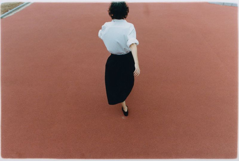 Woman in white shirt and black skirt walking on a terracotta-colored surface, captured in a wide shot from a high angle, minimalist.